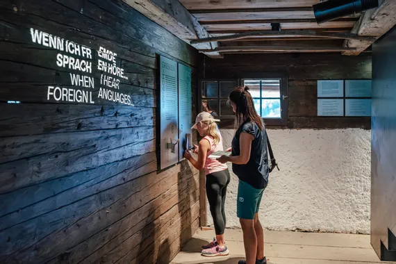 Zwei Besucherinnen interagieren mit einer mehrsprachigen Ausstellung in einem Holzraummuseum